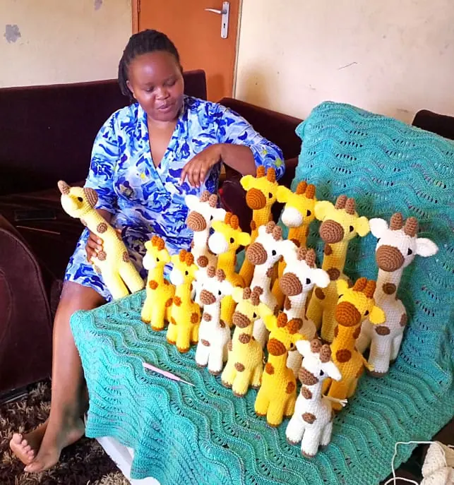 Woman in a blue patterned dress seated on a sofa beside many crocheted giraffes on a teal blanket