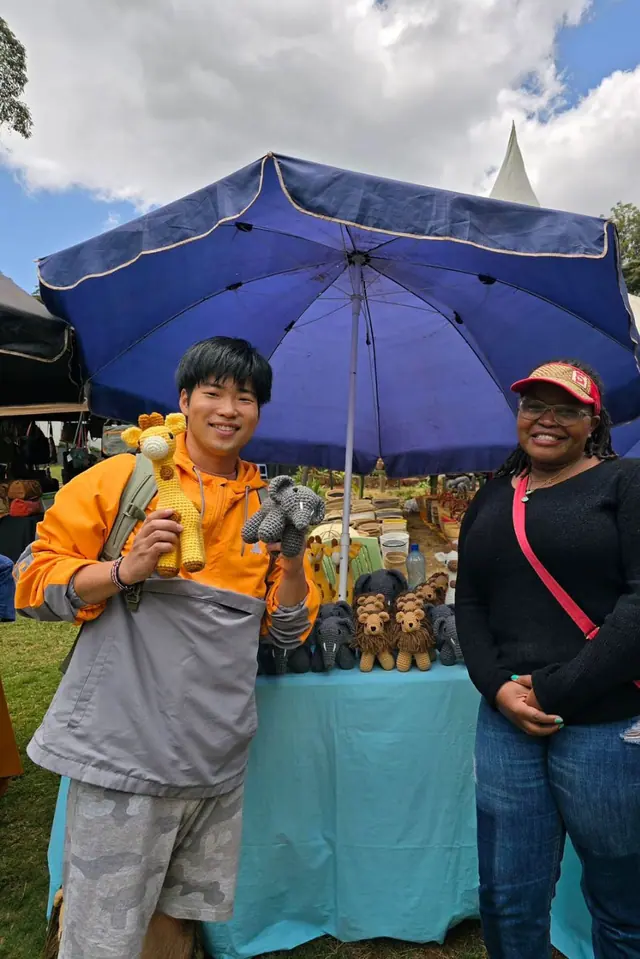 Young man holding a crocheted giraffe and elephant beside a smiling woman in a red visor under a blue market umbrella