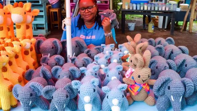 Woman with locs and glasses smiling behind a market table full of grey and blue crocheted elephants, rhinos and orange giraffes