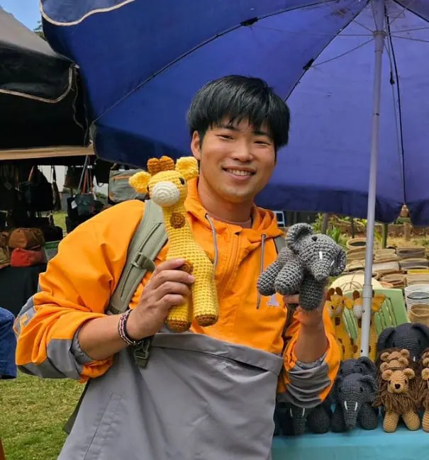 Smiling young man in an orange jacket holding a crocheted giraffe and a grey elephant under a blue umbrella